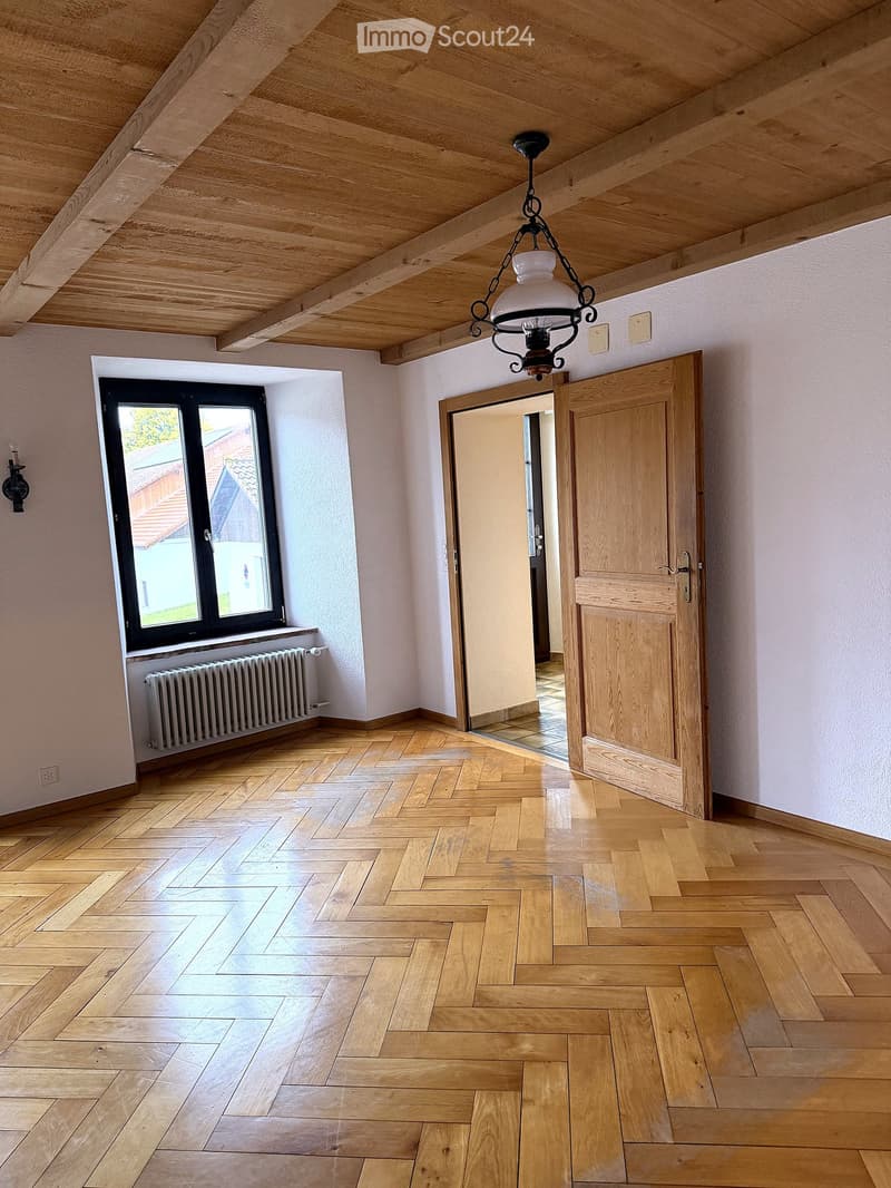 Empty room with wooden herringbone flooring, white walls, exposed wooden ceiling beams, a large window with black frame, a radiator, and a wooden door leading to another room.