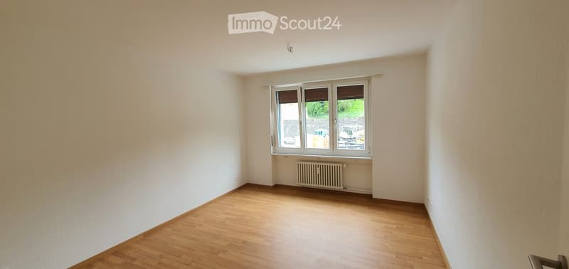 Empty room with wooden flooring, white walls, and a large window with blinds. A radiator is visible below the window.