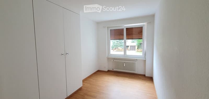 Empty room with wooden flooring, white walls, a large window with blinds, and a radiator below the window. A white closet is visible on the left side.