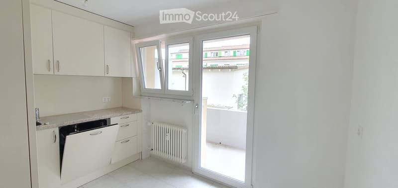 Modern kitchen with white cabinets, a dishwasher, and a radiator, featuring large windows and a sliding glass door leading to a balcony with a view of the city.
