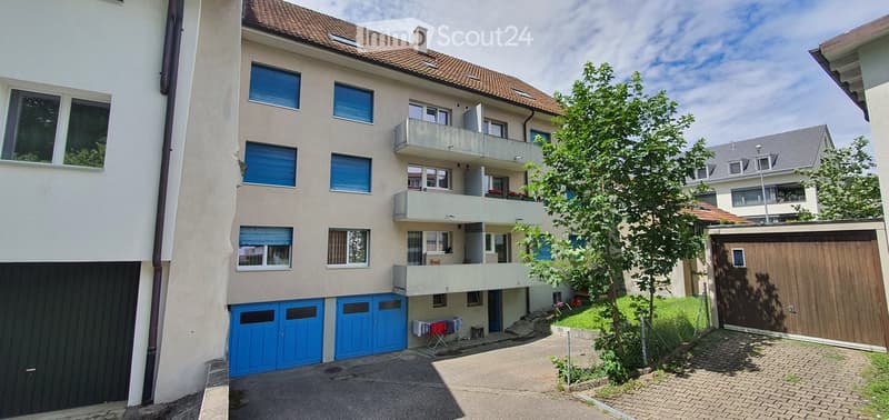 Multi-story residential building with balconies, blue garage doors, and a paved driveway.