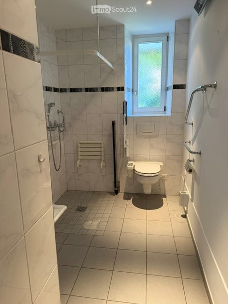 Modern bathroom with white tiled walls and floor, a toilet, a shower area with a handheld showerhead, a shower chair, and a towel rack with a handrail.