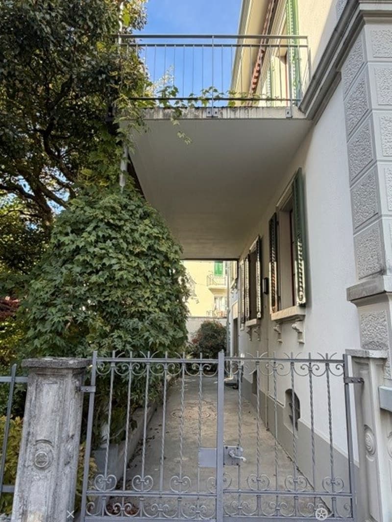 A multi-story residential building with a balcony accessible via a gated entrance. The building features classic architectural details, including ornate metalwork on the gate and balcony railing, and is surrounded by lush greenery.