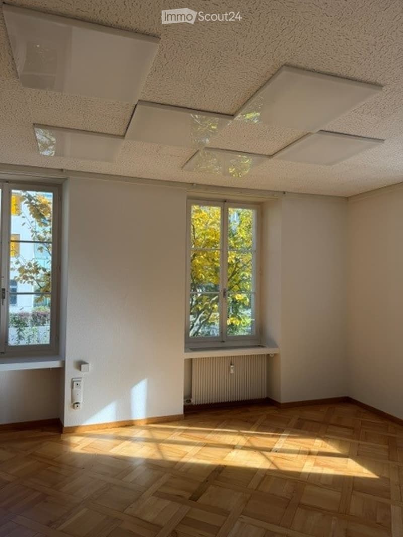 Empty room with wooden flooring, white walls, and large windows allowing natural light. No furniture or equipment present.