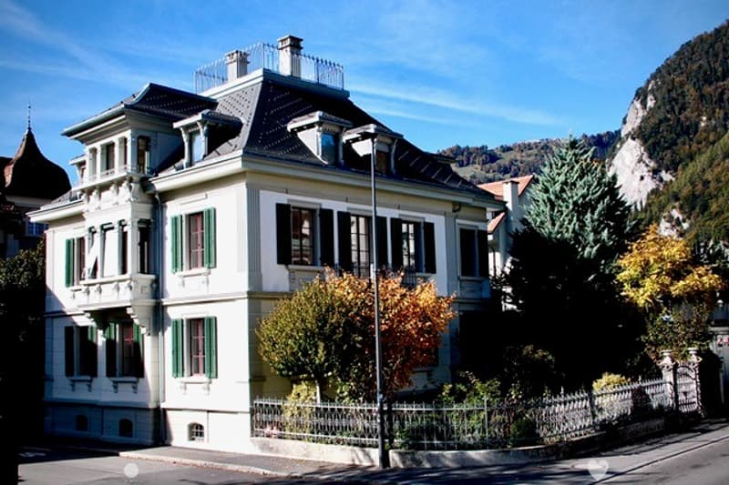 A large, multi-story house with a classic European architectural style, featuring white walls, green shutters, and a dark roof. The house is surrounded by a well-maintained garden with trees and a metal fence.