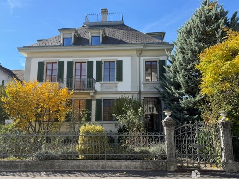A large, elegant two-story house with white walls, green shutters, and a grey roof. The house features multiple windows, a small balcony, and is surrounded by a well-maintained garden with trees and shrubs.