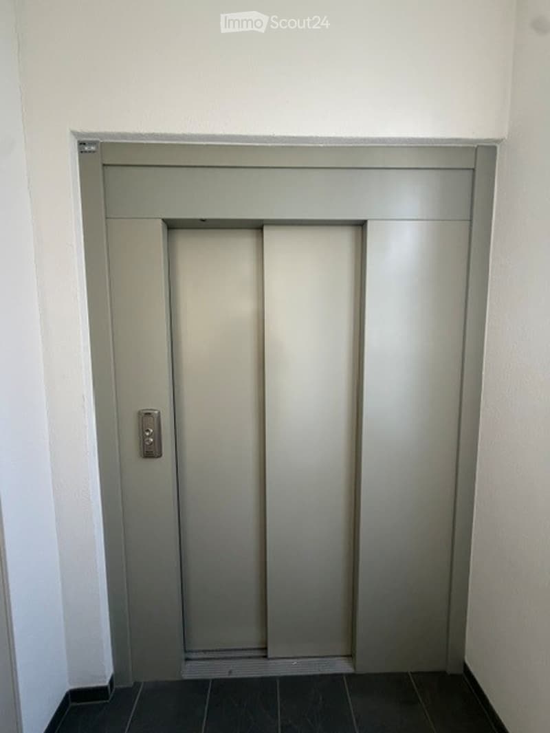 Elevator door in a hallway with a modern, minimalist design. The door is closed, featuring a sleek grey finish and a metallic handle.