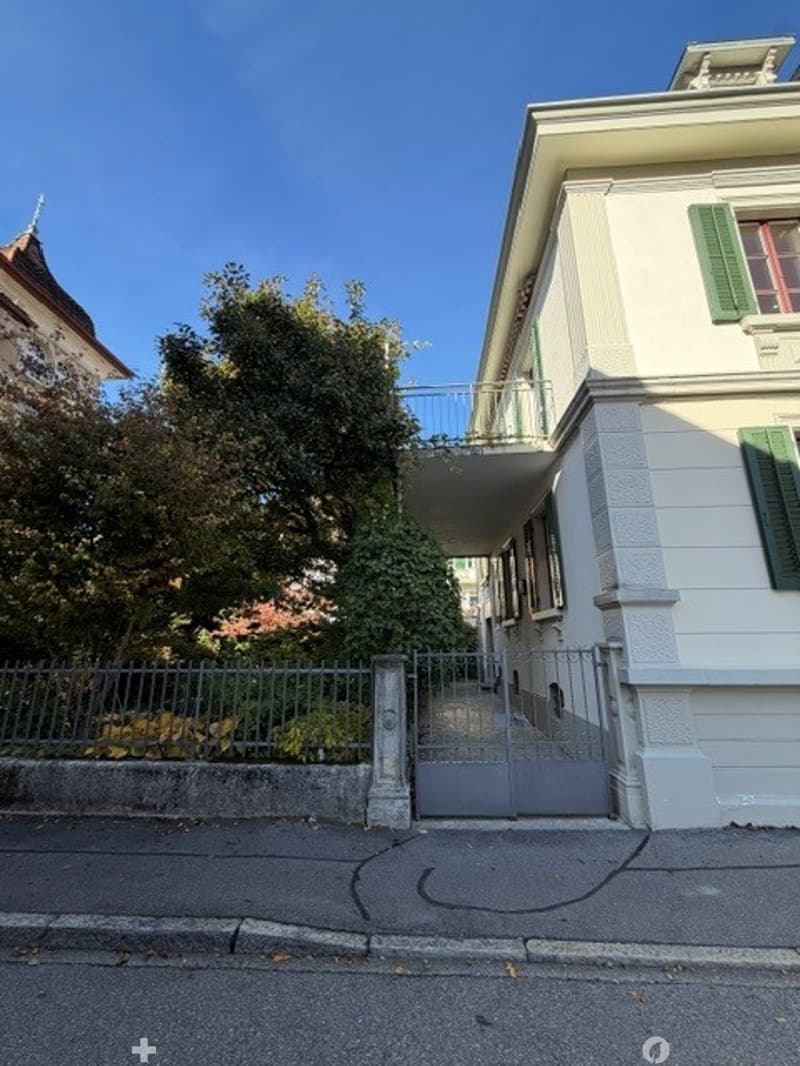 A two-story residential building with white walls, green shutters, and a balcony accessible from the upper floor. The entrance features a metal gate and is surrounded by a low fence and greenery.