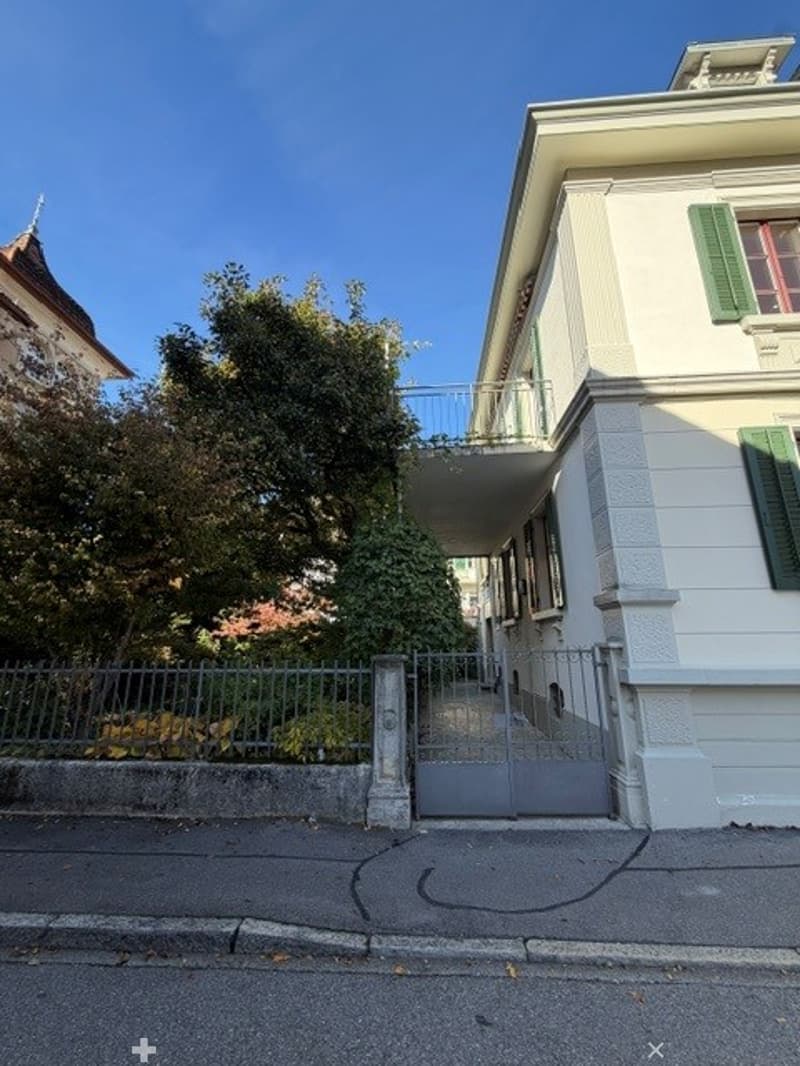 A two-story residential building with white walls, green shutters, and a balcony accessible from the upper floor. The entrance features a metal gate and is surrounded by a low fence and greenery.