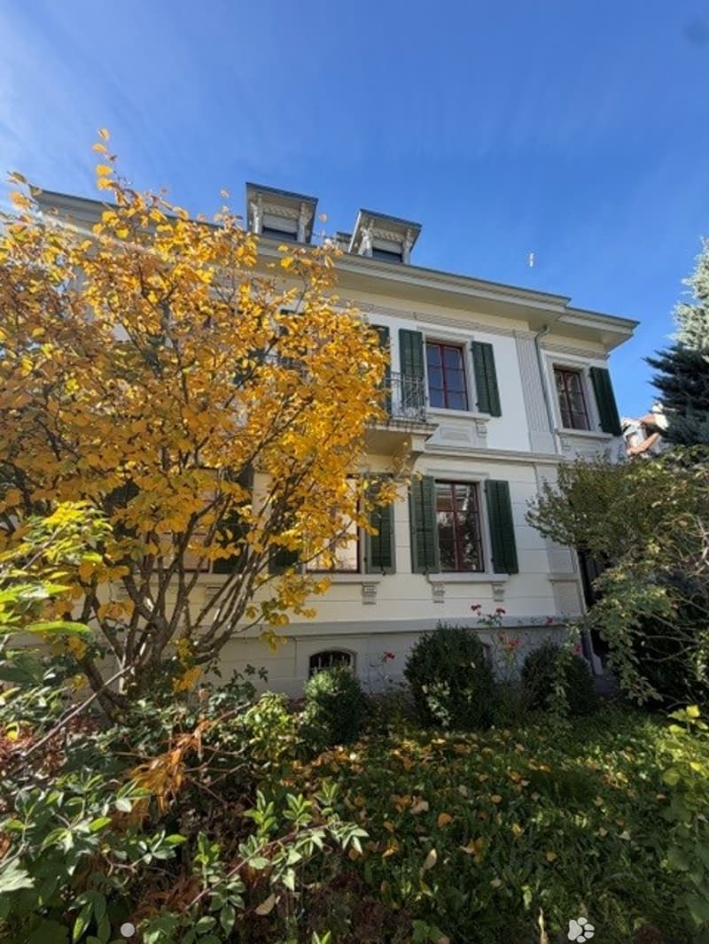 A multi-story residential house with white walls, green shutters, and multiple windows, surrounded by well-maintained greenery and trees.
