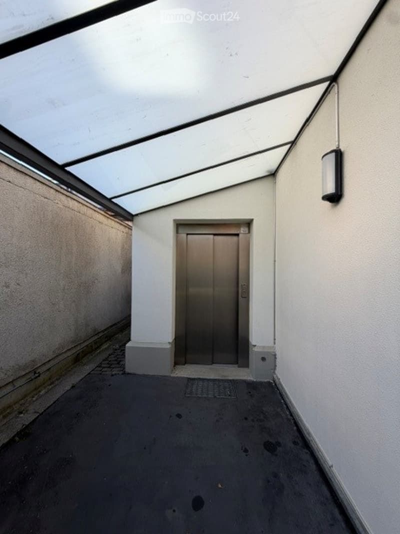 A narrow, covered corridor leading to an elevator door, with white walls and a gray floor.