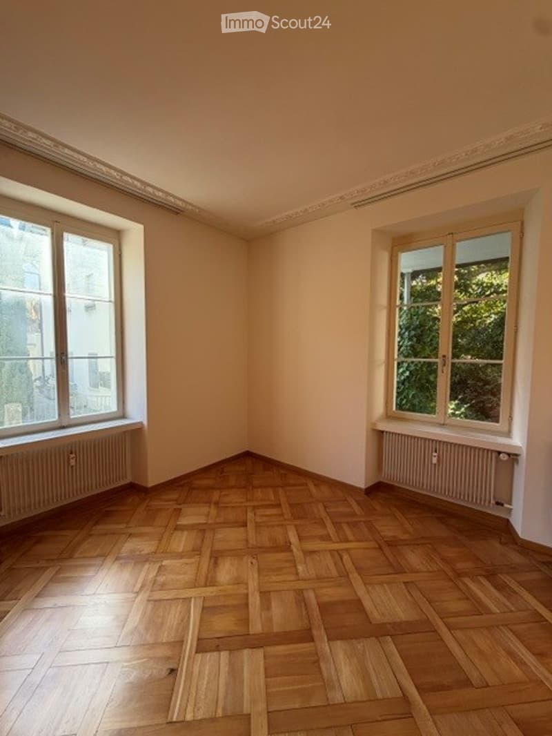 Empty room with wooden parquet flooring, two large windows, and radiators under the windows. The walls are painted a light beige color, and the ceiling has a decorative molding.