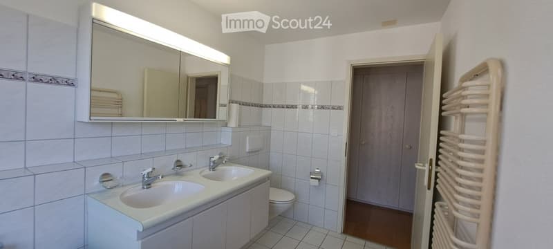 Modern bathroom with two sinks, a large mirror, a toilet, and a towel rack. The walls are tiled in light blue with a decorative border, and the floor is tiled in a matching pattern.