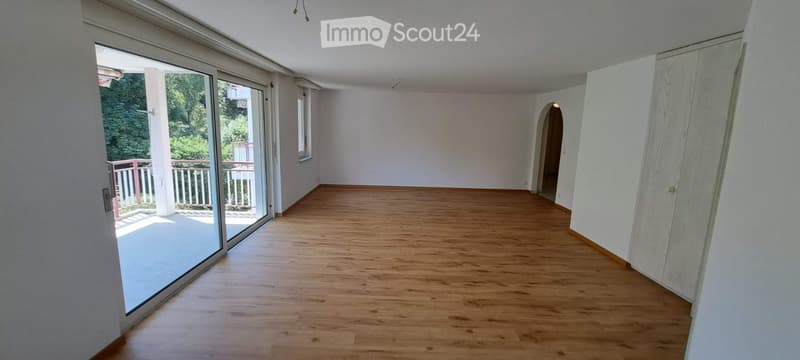 Empty room with wooden flooring, white walls, large sliding glass doors leading to a balcony, and a small arched doorway.