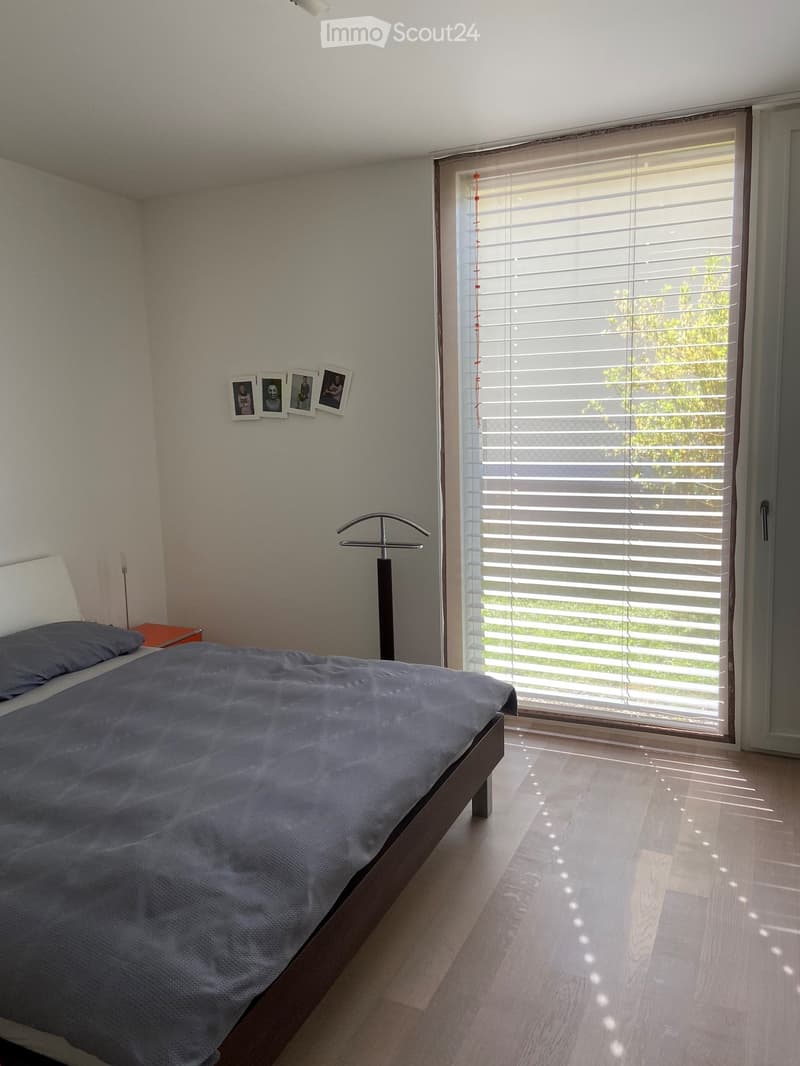 Modern bedroom with a wooden bed, nightstand, floor lamp, and picture frames on the wall. Large window with blinds allows natural light to enter.