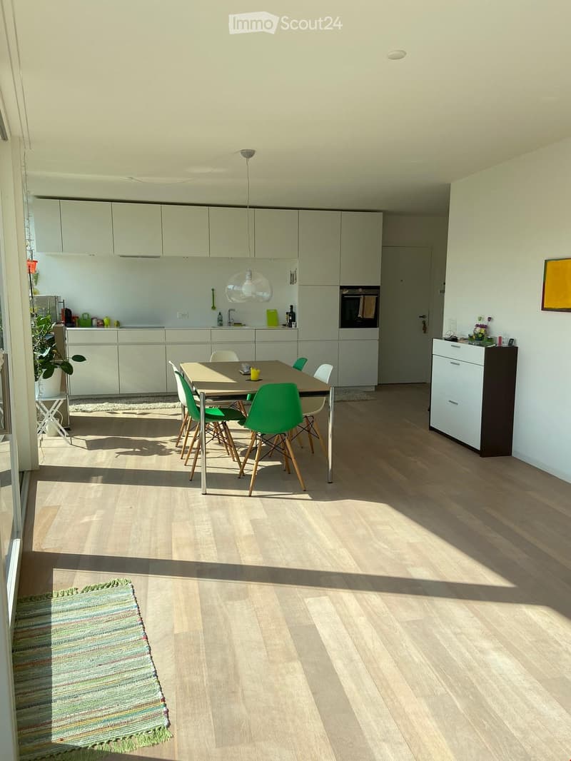 Modern kitchen with light-colored cabinetry, a central dining table with green chairs, pendant lighting, and a small plant near the window.