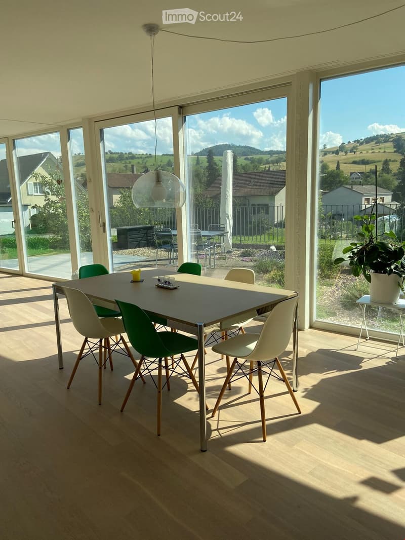 Bright, modern kitchen with large windows offering a scenic view, featuring a dining table with chairs, a potted plant, and a candle on the table.