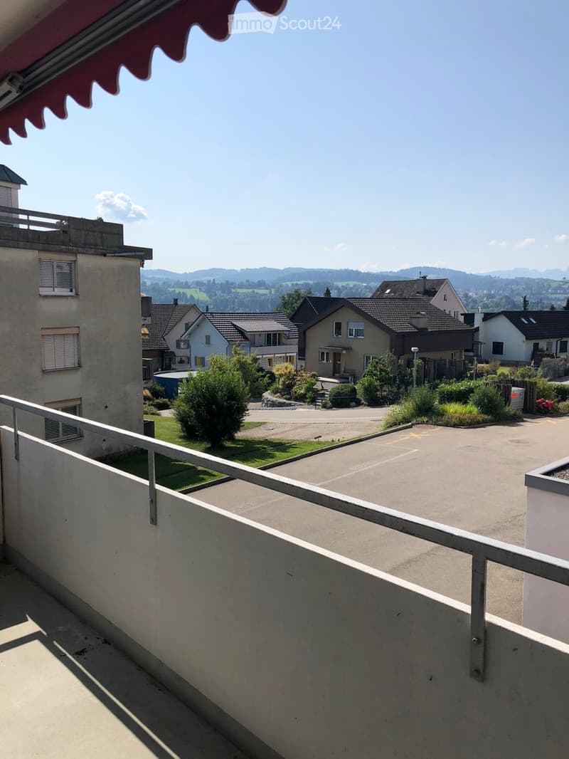 Balcony overlooking a residential area with houses, greenery, and a clear view of distant hills under a bright blue sky.