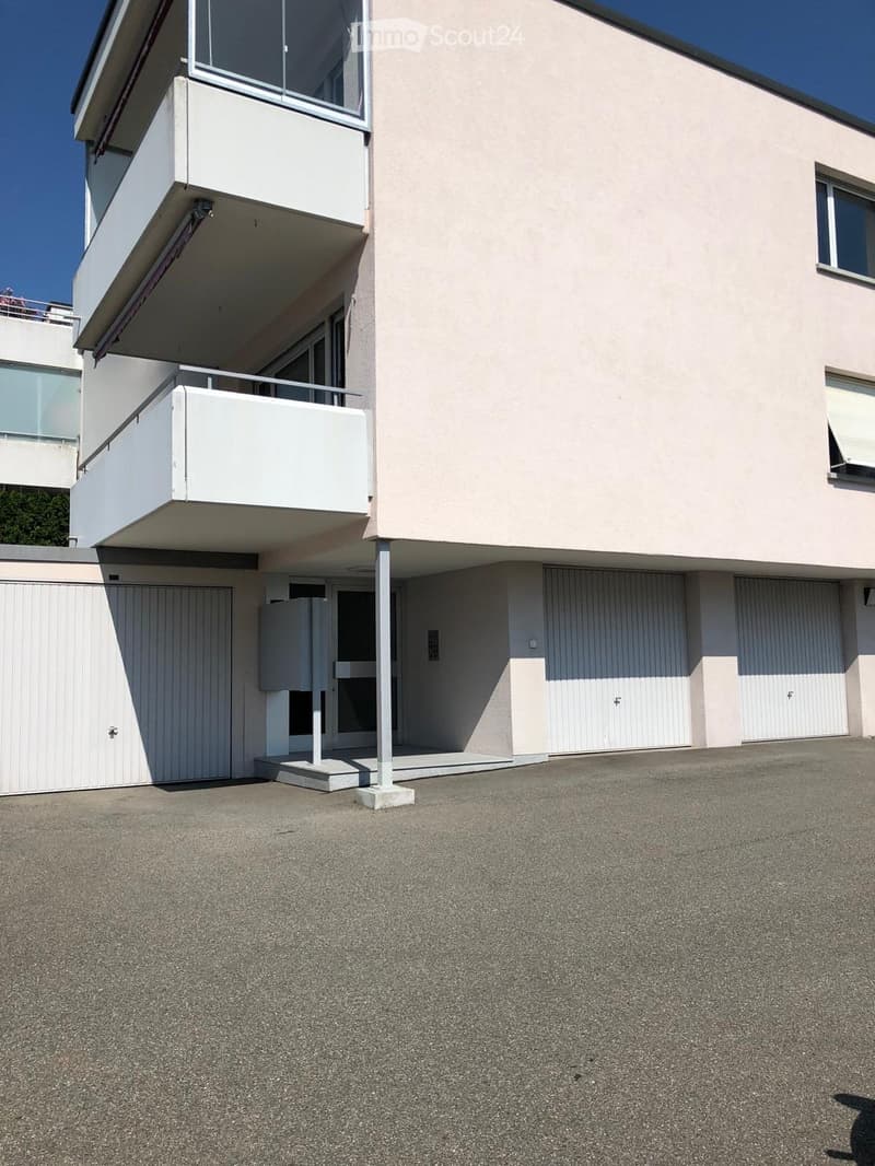 Modern two-story building with light pink facade, large windows, and balconies. Features include a raised ground floor, garage, and parking area.