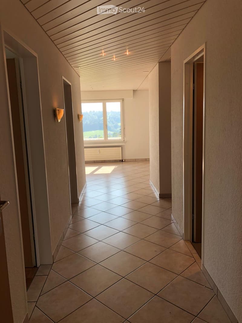 Empty hallway with tiled floor, beige walls, and a large window at the end showing a scenic view. The ceiling has recessed lighting and a wooden panel design.