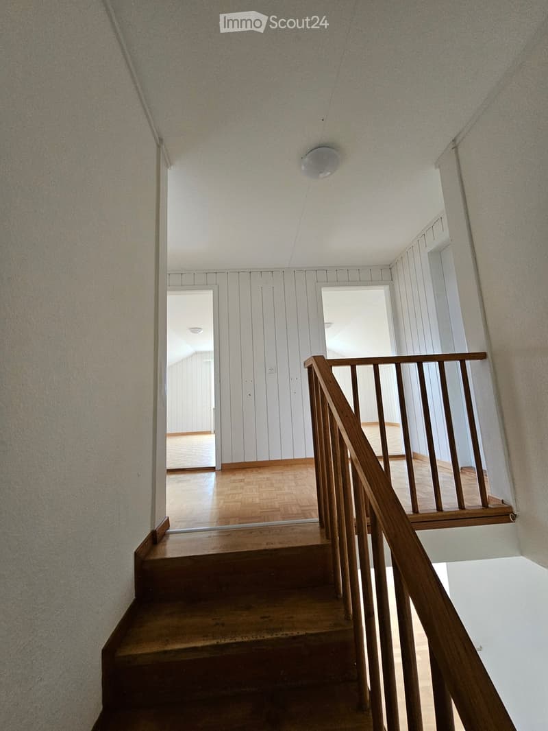 Wooden staircase with a wooden handrail leading to an open area with wooden flooring and white walls.