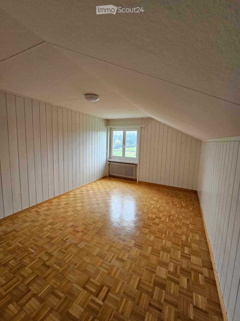Empty room with wooden herringbone flooring, white paneled walls, and a single window with a radiator below it.