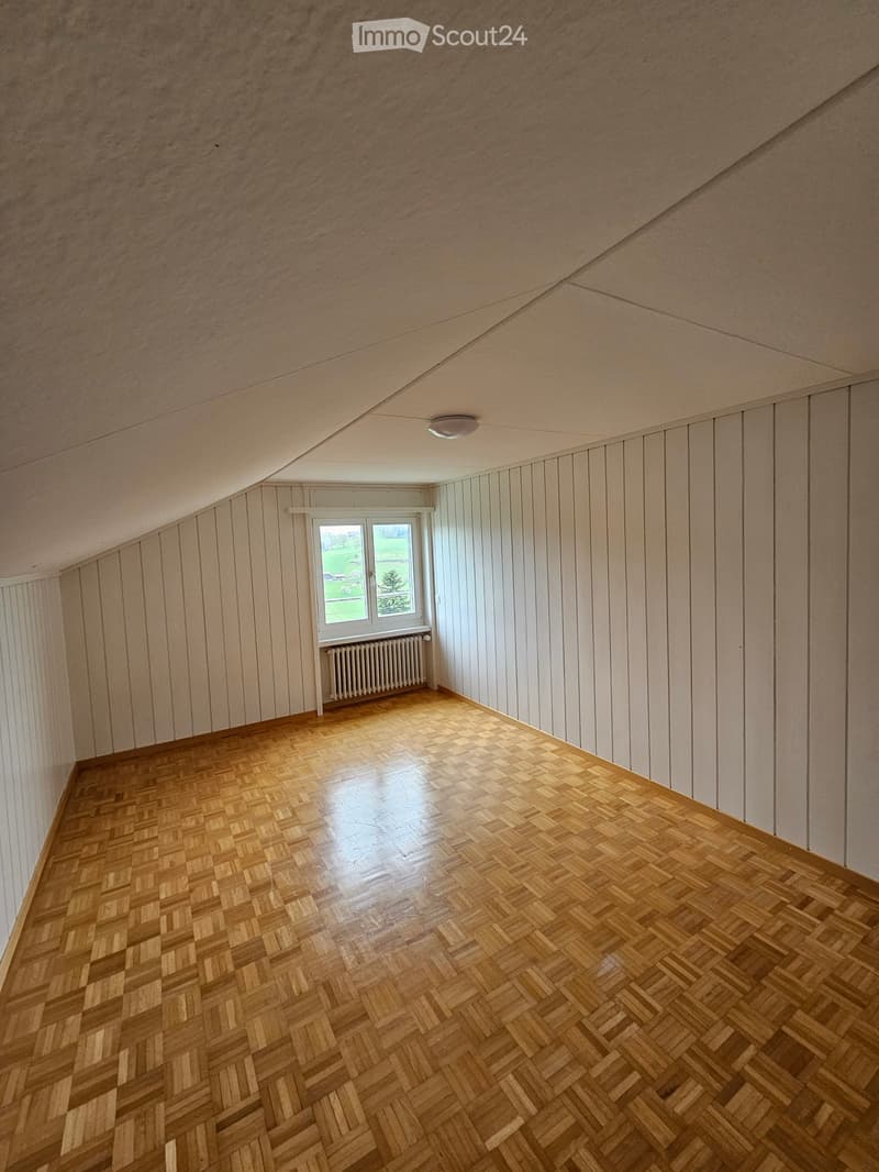 Empty room with wooden herringbone flooring, white paneled walls, and a single window with a radiator below it.