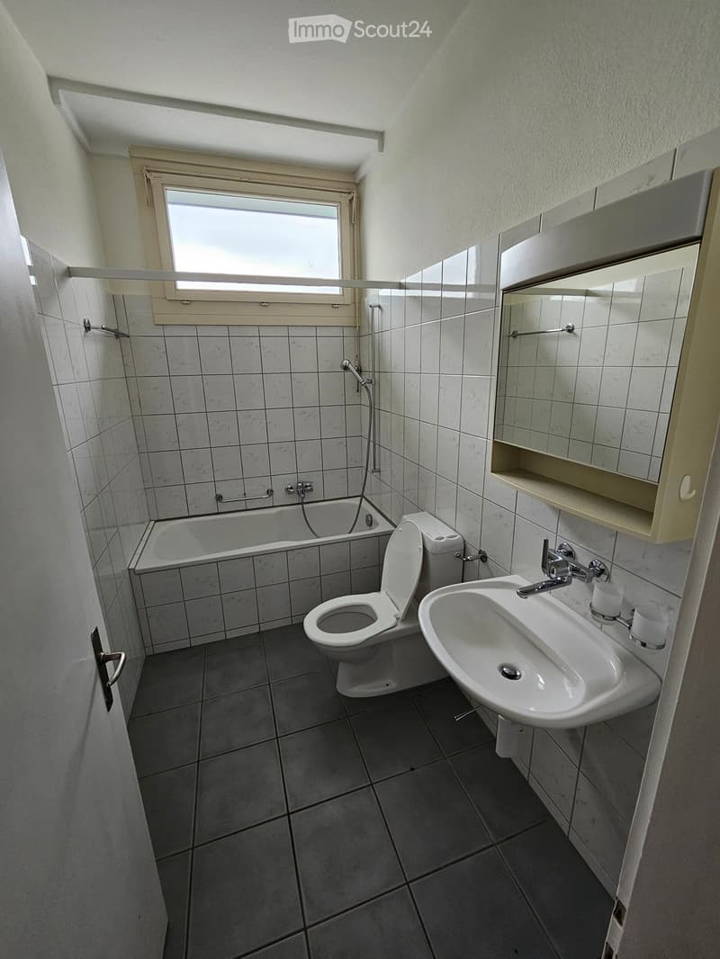 Bathroom with a bathtub, toilet, sink, mirror, and cabinet. The room features tiled walls and a window above the bathtub.