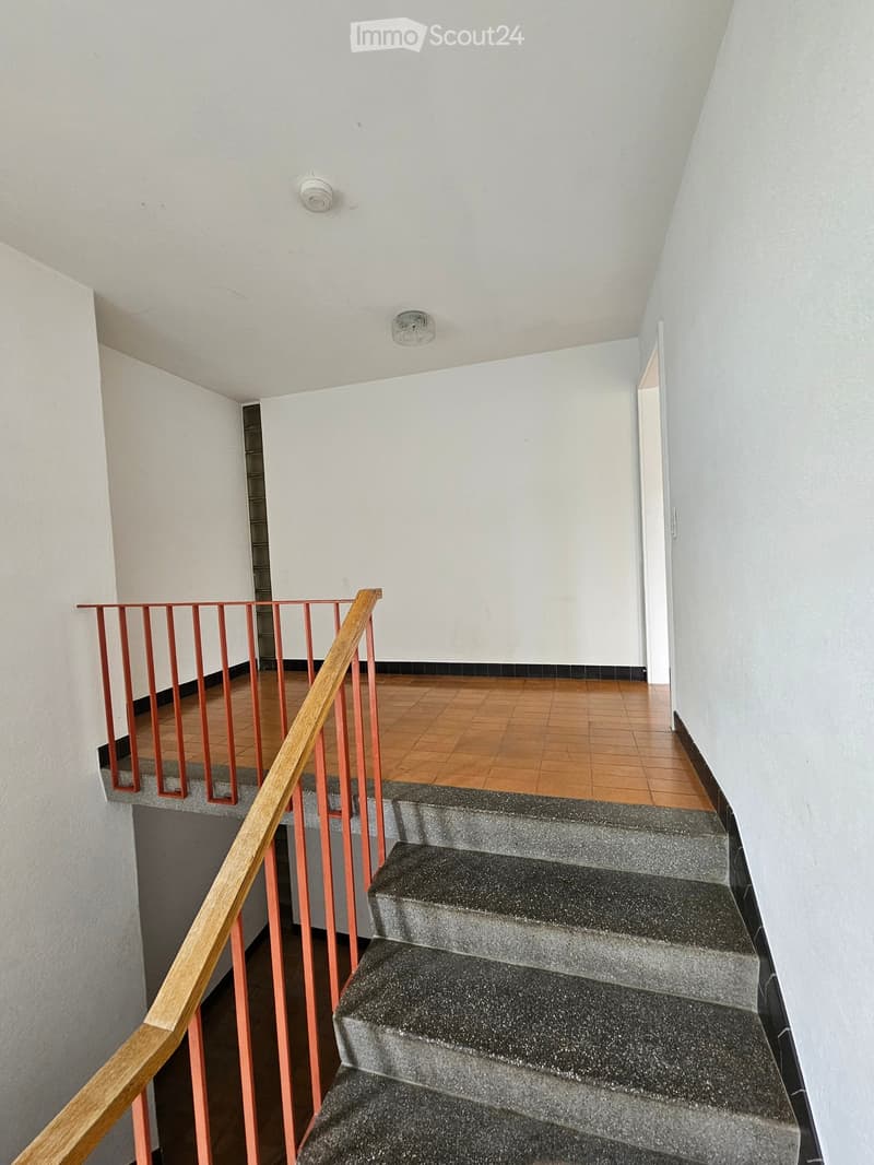 Staircase with wooden handrail and red metal balustrade, leading to an open area with wooden flooring and white walls.