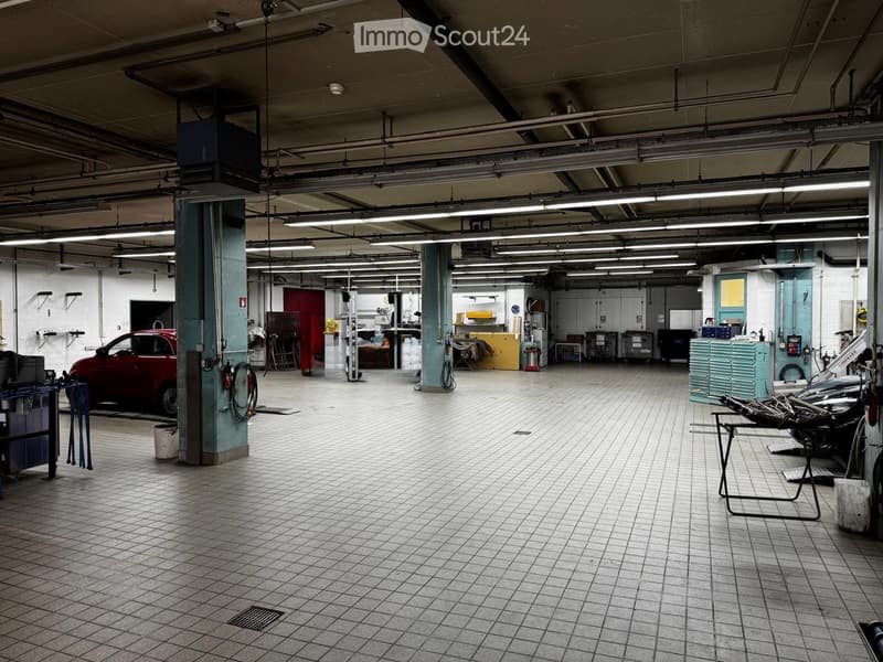 A spacious garage with tiled flooring, fluorescent lighting, and various automotive tools and equipment. A red car is parked on the left, and workbenches and storage units are visible throughout the space.