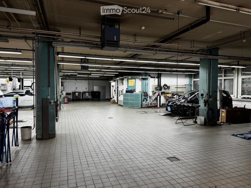 A spacious, well-lit garage with tiled flooring, industrial lighting, and various tools and equipment. Several cars are parked inside, and there are cabinets and storage units along the walls.