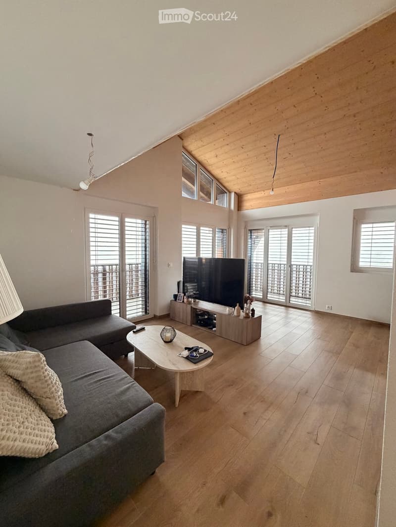 Modern living room with wooden flooring, large windows, and a balcony. Features a grey sectional sofa, a white coffee table, a television, and two lamps.