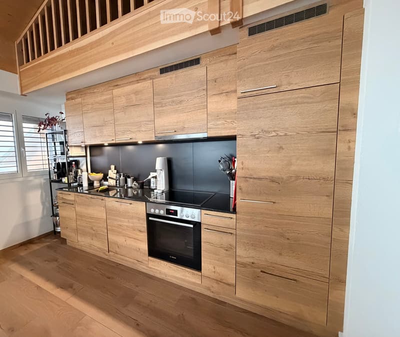 Modern kitchen with light wooden cabinets, stainless steel appliances, and a black countertop. Features include an oven, coffee maker, and various kitchen utensils.