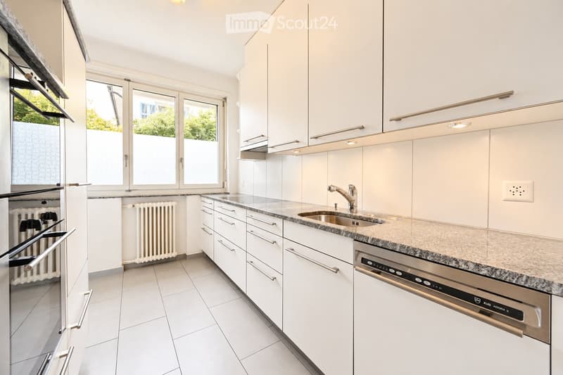 Modern kitchen with white cabinets, granite countertop, stainless steel sink and dishwasher, large windows, and tiled floor.
