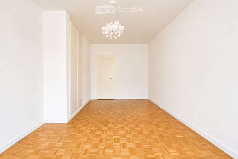 Empty hallway with wooden herringbone flooring, white walls, and a single hanging light fixture.