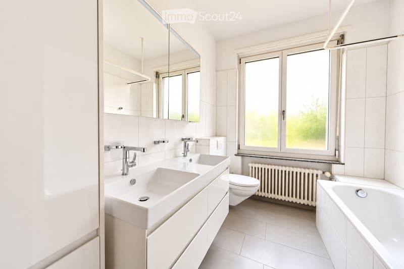Modern bathroom with white tiles, double sink, large mirror, toilet, bathtub, and radiator under a window.