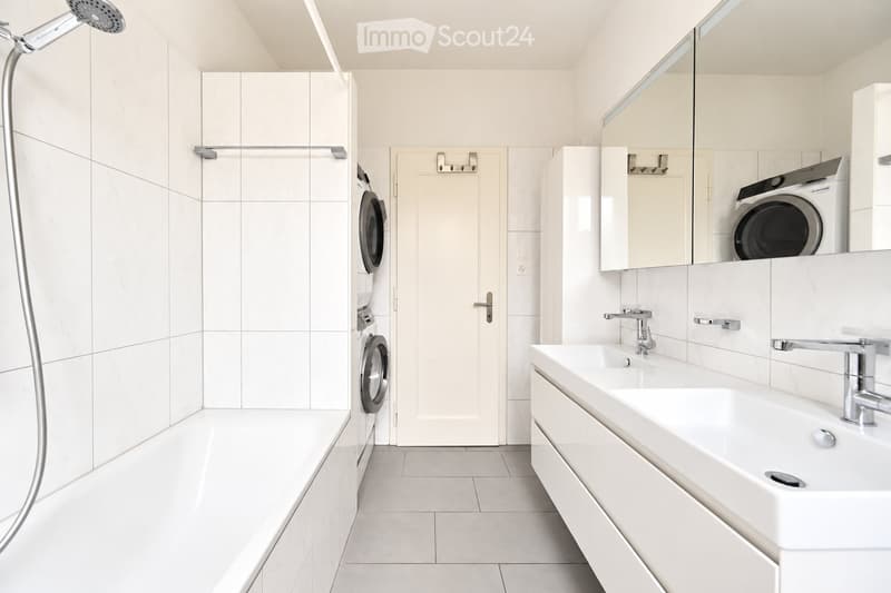 Modern bathroom with white tiles, a bathtub, a sink with a mirror, a washing machine, and a showerhead.