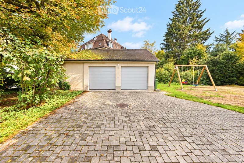 A paved driveway leads to a two-car garage with white doors, surrounded by greenery and a swing set in the background.