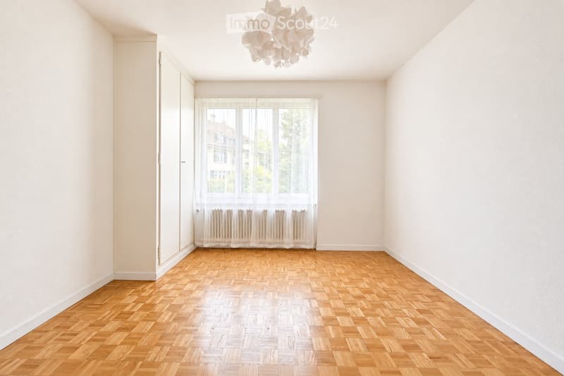 Empty room with wooden herringbone flooring, white walls, large window with sheer curtains, and a modern white chandelier.