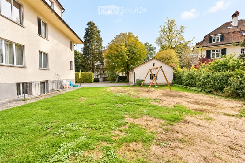 A well-maintained garden with a mix of grass and sandy areas, a swing set, and surrounding trees and houses.