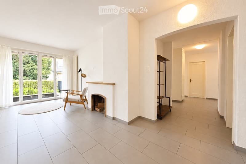 Bright living room with white walls, tiled floor, fireplace, white chair, small table, and a hallway leading to another room.