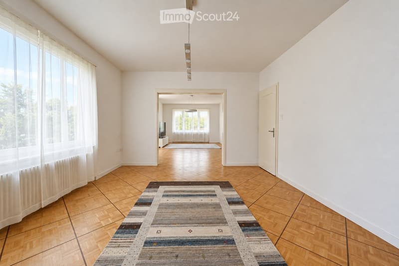 Bright hallway with wooden flooring, large windows with sheer curtains, a patterned rug, and a pendant light fixture. An open doorway leads to another room with a TV and additional windows.