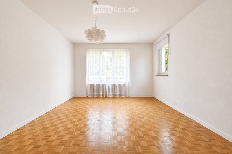 Empty room with wooden parquet flooring, white walls, large windows with sheer curtains, and a modern hanging light fixture.
