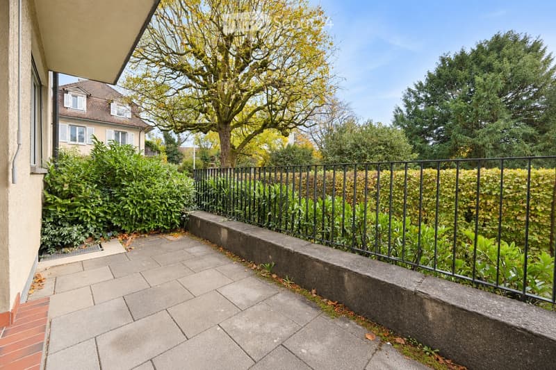 Balcony with tiled floor, black metal railing, and surrounding greenery including bushes and trees.