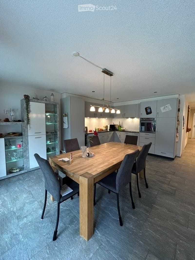 Modern kitchen with a wooden dining table surrounded by six chairs, grey cabinets, a refrigerator, and pendant lights above the island.