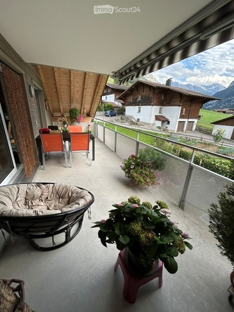 Balcony with a view of the mountains, featuring a round wicker chair, a small table with a flower pot, and a couple of orange chairs with a small table.