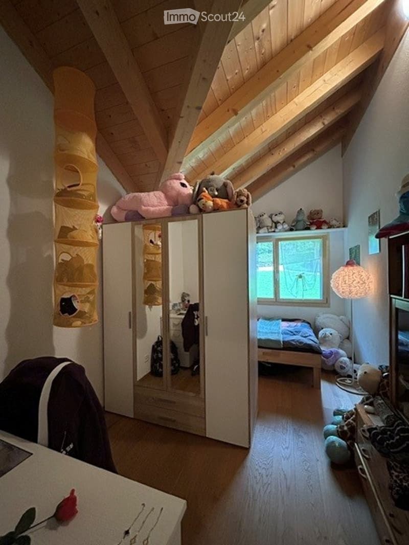 Cozy children's bedroom with wooden slanted ceilings, a large wardrobe, a bed with stuffed toys, and a hanging storage basket.