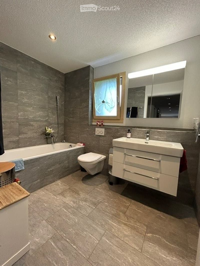 Modern bathroom with a bathtub, toilet, sink, and large mirror. Features include a grey tiled wall, a window with a sheer curtain, and a light fixture above the mirror.