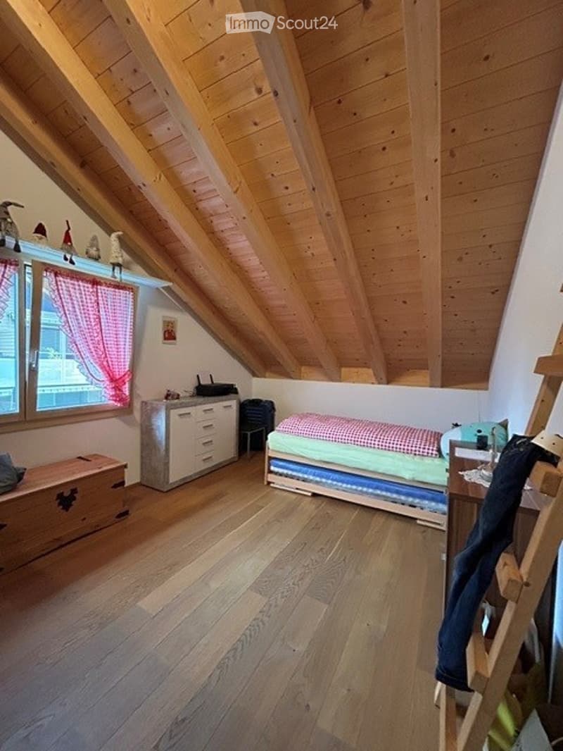 Cozy attic bedroom with wooden slanted ceilings, hardwood floors, a single bed with colorful bedding, a small cabinet, and a window with red curtains.