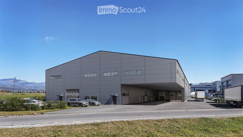 Large industrial building, gray exterior, large doors, multiple windows, flat roof, vehicles parked outside